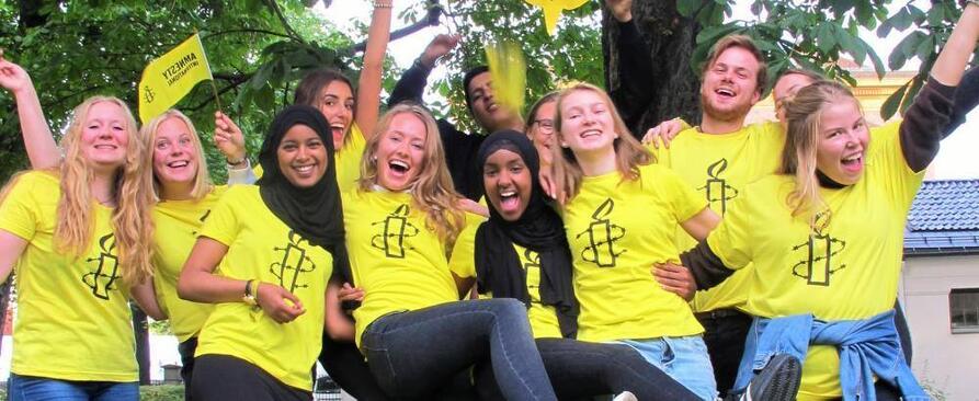 En gruppe jenter og gutter i gule Amnesty T-skjorter med Amnestys logo på, jubler og rekker armene i været. De har gule Amnesty-flagg som de vifter med. Noen av jentene har langt lyst hår, guttene har kort lyst eller mørkt hår og to av jentene har svart hijab.