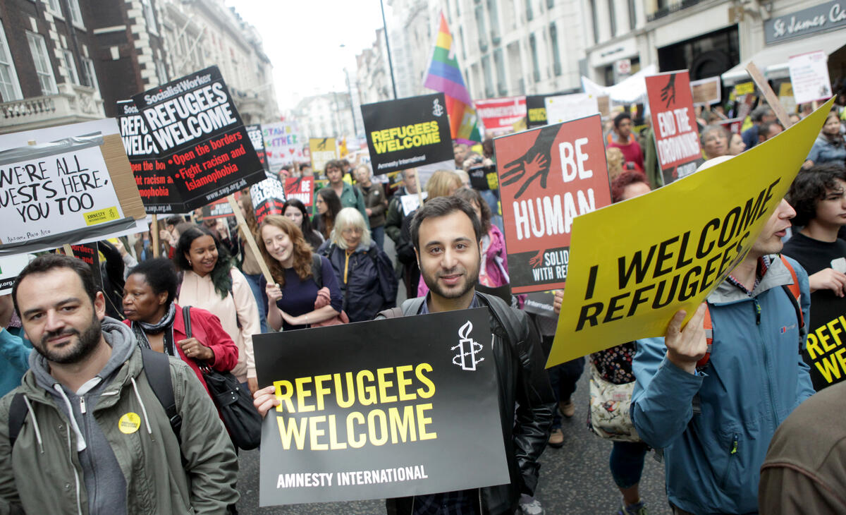 Mennesker går i parade med plakater. Teksten på plakaten i fokus sier "Welcome refugees".