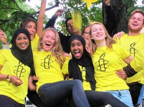 En gruppe jenter og gutter i gule Amnesty T-skjorter med Amnestys logo på, jubler og rekker armene i været. De har gule Amnesty-flagg som de vifter med. Noen av jentene har langt lyst hår, guttene har kort lyst eller mørkt hår og to av jentene har svart hijab.